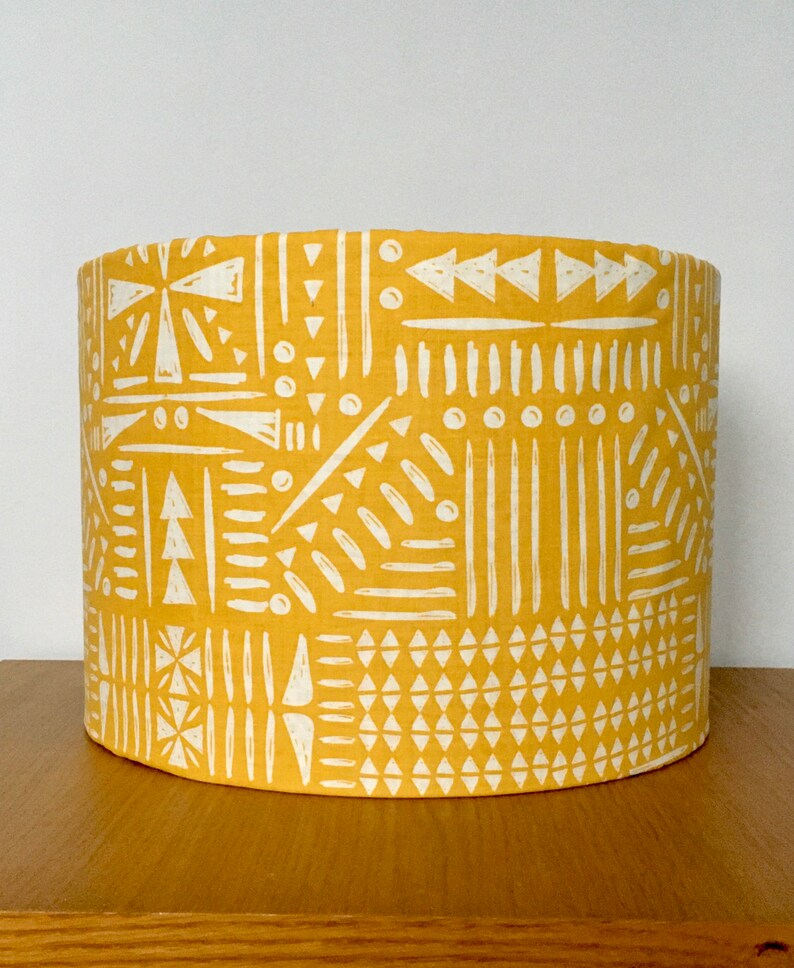 May include: A cylindrical lampshade featuring a vibrant mustard yellow background with a repeating white geometric pattern. The design consists of various hand-drawn style shapes including triangles, vertical lines, dots, and dashes arranged in panels.
