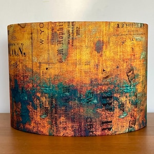 May include: Cylindrical lampshade with a vintage newspaper print design. The shade features a blend of orange, yellow, and teal hues, with visible text fragments. The lampshade is designed to be placed on a table or other surface.