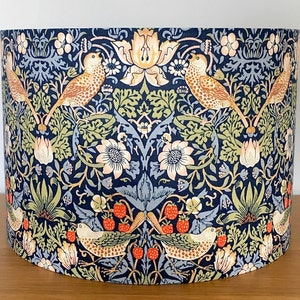 May include: A fabric lampshade featuring a repeating pattern of birds, flowers, and foliage in beige, green, and orange on a dark blue background. The lampshade has a cylindrical shape and is designed for a lamp base.