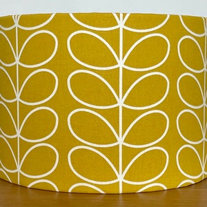 May include: Mustard yellow lampshade with a white leaf pattern. The cylindrical lampshade is made of fabric and features a repeating design of white, stylized leaves against a solid yellow background. The lampshade is designed to fit a standard lamp base.