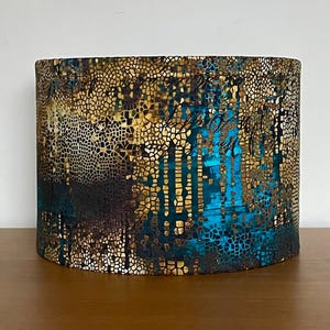 May include: A decorative lampshade with a mosaic design in gold, teal, and dark brown. The cylindrical lampshade has a textured surface with a mix of colours and shapes, creating a unique lighting feature.