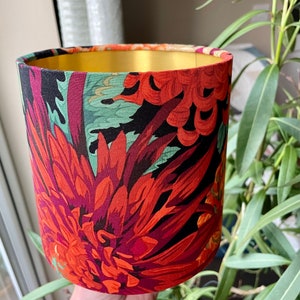 Op de afbeelding: Een rode, groene en zwarte bloemenlampkap met een gouden bovenkant. De lampkap heeft een grote rode bloem met groene bladeren.