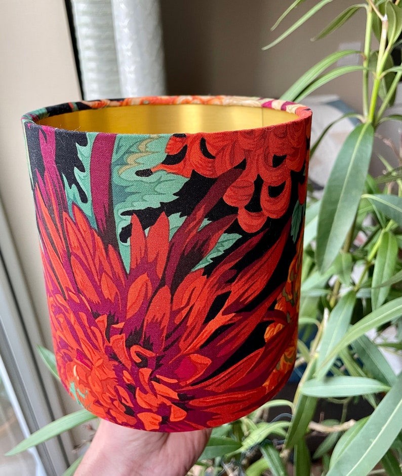 Op de afbeelding: Een rode, groene en zwarte bloemenlampkap met een gouden bovenkant. De lampkap heeft een grote rode bloem met groene bladeren.