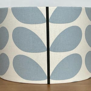 May include: Cylindrical lampshade with a cream background and a repeating pattern of light blue leaf shapes. A vertical black stripe bisects the shade. The lampshade is sitting on a wooden surface.