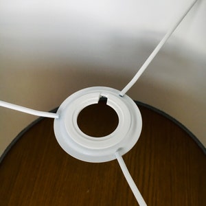 May include: A white metal lamp shade fitter with a circular center ring and three radiating support arms. The fitter is attached to the interior of a lamp shade, featuring a smooth white finish on the metal components and a dark brown interior.