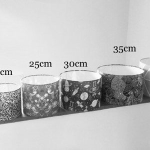 May include: A row of six cylindrical lampshades in varying sizes, labeled 15cm, 20cm, 25cm, 30cm, 35cm, and 40cm. The shades feature diverse patterns including floral, space-themed, and geometric designs in black, white, and gray tones with white interiors.