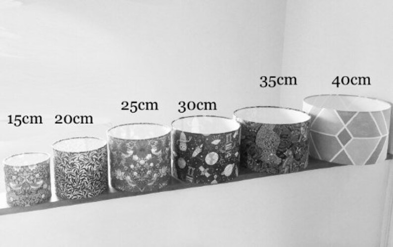 May include: A row of six cylindrical lampshades in varying sizes, labeled 15cm, 20cm, 25cm, 30cm, 35cm, and 40cm. The shades feature diverse patterns including floral, space-themed, and geometric designs in black, white, and gray tones with white interiors.