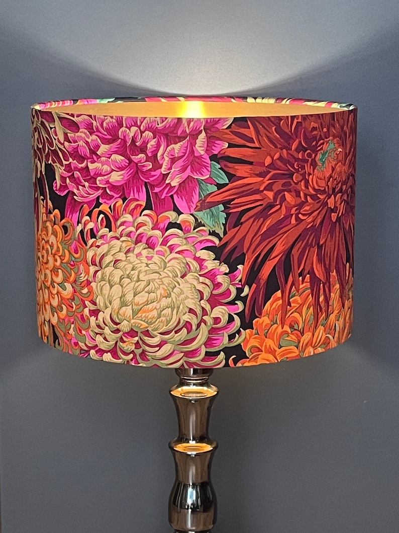 Op de afbeelding: Een gouden tafellamp met een zwart-witte bloemenlampkap met roze, oranje en rode chrysanten.