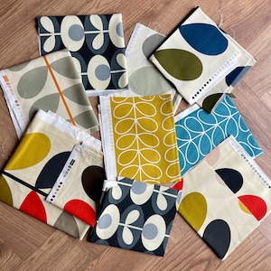 May include: A selection of fabric samples showcasing diverse abstract patterns. The swatches display designs in colours such as mustard yellow, teal, navy, and olive green. Some patterns feature leaf-like shapes and geometric designs. The fabrics are arranged on a wooden surface.