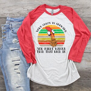 May include: A white and red baseball tee shirt with a retro-style graphic of a chicken with the text "Your crazy is showing. You might wanna tuck that back in."