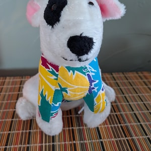 May include: A white plush dog with black spots and pink ears, wearing a colorful shirt. The shirt features a tropical leaf pattern in yellow, blue, green, and purple. The toy is sitting on a brown and tan bamboo mat.