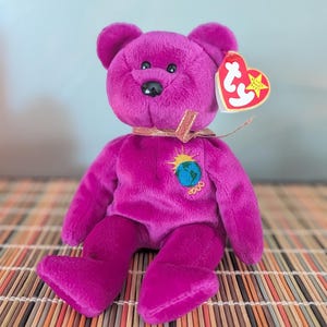 May include: A magenta-coloured plush bear with a black nose and small, round eyes. The bear has a gold ribbon around its neck and a patch on its chest with a globe and sun design. A red heart-shaped tag is attached to its ear.