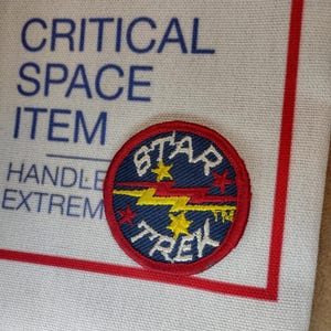 May include: A circular Star Trek patch with a red border, blue center, and white lettering. The patch features a yellow lightning bolt and stars. The background includes text that reads "CRITICAL SPACE ITEM HANDLE EXTREM".