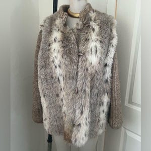 Pode incluir: Um colete de pele sintética com um padrão salpicado em tons de cinza e branco, combinado com mangas de malha. O colete tem gola e fecha com dois botões. A peça de vestuário é exibida num manequim.
