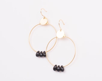 May include: A pair of gold-colored hoop earrings with a small, round gold disc at the top and three black teardrop-shaped beads hanging from the bottom of each hoop. The earrings are set against a white background.