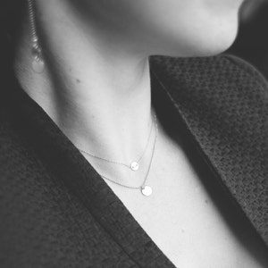 Peut inclure: Un collier en argent avec deux chaînes délicates et deux pendentifs ronds. Le collier est porté par une personne avec une veste sombre.