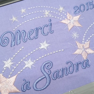 Peut inclure: Un tissu violet clair avec un message brodé en bleu qui dit "Merci a Sandra" avec une étoile rose et des étoiles blanches et un fil de perles blanches. L'année 2015 est brodée dans le coin supérieur droit.