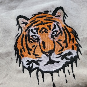 Puede incluir: Diseño de cara de tigre bordada sobre un fondo de tela blanca. El tigre tiene rayas naranjas y negras y un hocico blanco.