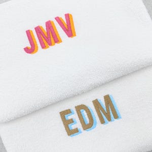 May include: Two white towels with embroidered initials. The top towel features the initials "JMV" in pink and orange, while the bottom towel displays "EDM" in brown and blue. The towels are folded and stacked.