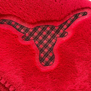 May include: Close-up of a red, plush textile featuring an embroidered longhorn silhouette. The longhorn is filled with a black and red plaid pattern. The background is a soft, textured red.