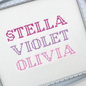 Puede incluir: Bordado de punto de cruz con los nombres STELLA, VIOLET y OLIVIA en tonos rosas y morados. Los nombres están bordados sobre tela blanca, enmarcados por un bastidor gris.