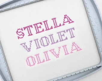 May include: Cross-stitch embroidery featuring the names STELLA, VIOLET, and OLIVIA in varying shades of pink and purple. The names are stitched on white fabric, framed by a gray embroidery hoop.
