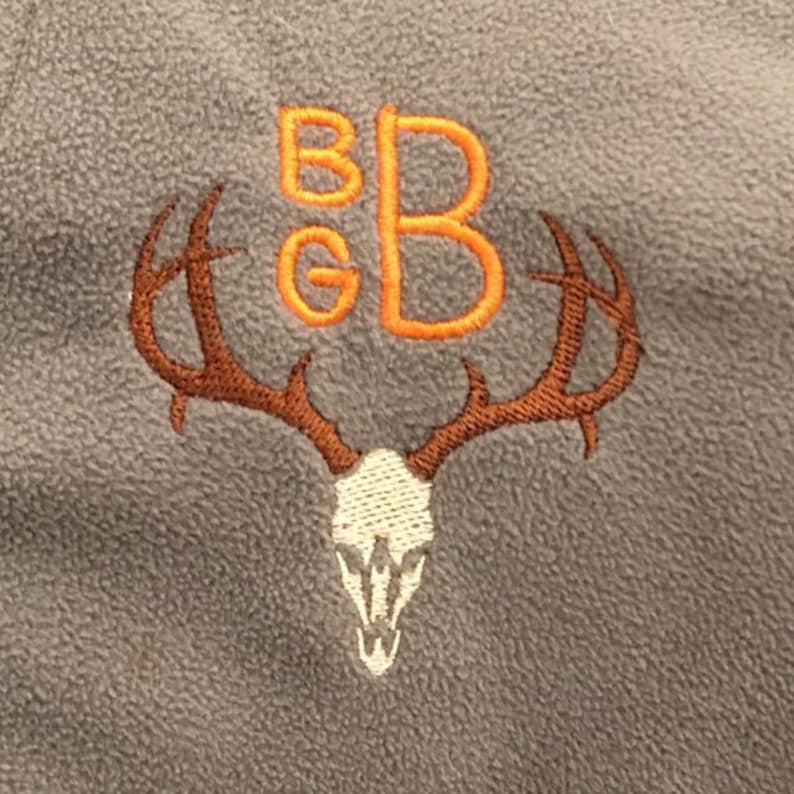 Puede incluir: Una tela de tejido polar gris con un cr&aacute;neo de ciervo y cuernos bordados en marr&oacute;n. Las letras "BGB" est&aacute;n bordadas en naranja sobre el cr&aacute;neo.