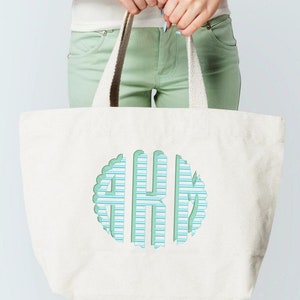 May include: A white canvas tote bag with a green and blue striped monogram in a circle. The monogram is the letters "AKM".