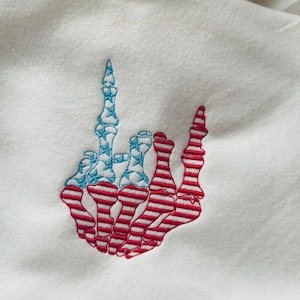 May include: Embroidered design of a skeleton hand giving the middle finger, with the bones patterned in the colors of the American flag.