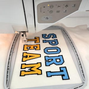 May include: A white embroidery machine with a digital control panel is stitching the words SPORT TEAM in block letters onto white fabric held in a gray hoop. The word SPORT is in light blue and TEAM is in yellow, both with black outlines.