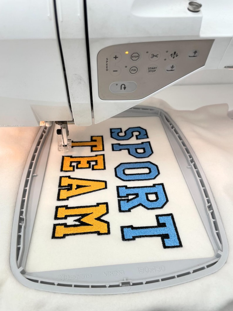 May include: A white embroidery machine with a digital control panel is stitching the words SPORT TEAM in block letters onto white fabric held in a gray hoop. The word SPORT is in light blue and TEAM is in yellow, both with black outlines.