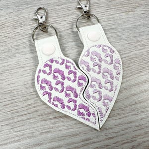 May include: Two white heart-shaped keychains with a split design, featuring purple embroidered leopard print patterns. Each keychain includes a silver-toned metal lobster clasp, a circular split ring, and a white snap button closure at the top.