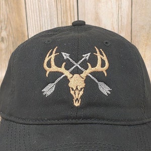 Puede incluir: Gorra de b&eacute;isbol negra con un dise&ntilde;o bordado de una calavera de ciervo con cuernos y dos flechas cruzadas. La calavera es dorada y las flechas son plateadas.