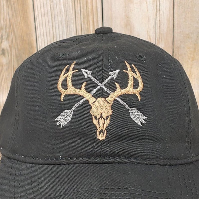 Puede incluir: Gorra de b&eacute;isbol negra con un dise&ntilde;o bordado de una calavera de ciervo con cuernos y dos flechas cruzadas. La calavera es dorada y las flechas son plateadas.