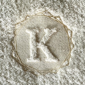 May include: A white towel with a beige embroidered monogram of the letter "K" in a circular design.