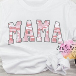 May include: White t-shirt with the word "MAMA" in pink and white camouflage lettering. The letters have a stitched outline.