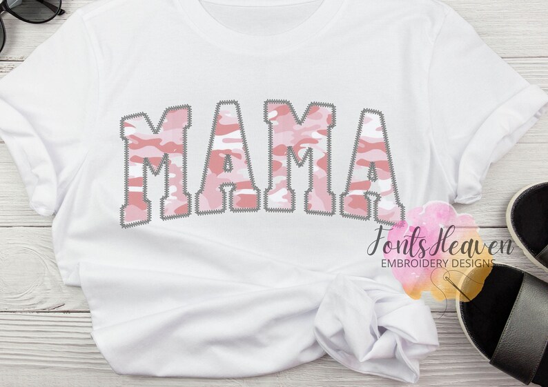 May include: White t-shirt with the word "MAMA" in pink and white camouflage lettering. The letters have a stitched outline.