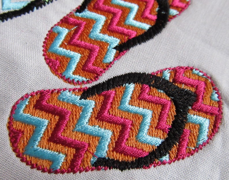 May include: Close-up of two embroidered flip-flop designs. The flip-flops are orange, pink, and blue with a black outline. The designs are stitched in a chevron pattern.