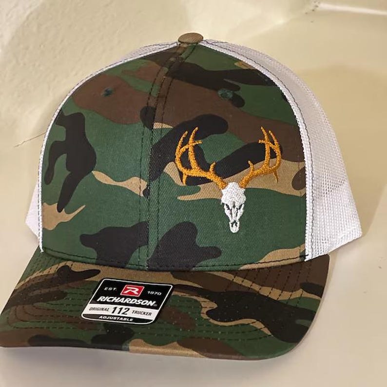 Puede incluir: Un gorro de camionero de camuflaje y blanco con una calavera de ciervo blanca y cuernos bordados en la parte delantera. El gorro tiene una correa ajustable y un logotipo de Richardson 112.