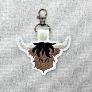 May include: A white and brown embroidered Highland cow keychain with a bronze-colored clasp. The keychain features a detailed design of a Highland cow's head with brown fur, black hair, and tan horns.