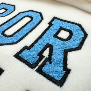 May include: A white fabric surface featuring embroidered lettering with light blue fill and thick black outlines. The visible text includes the letters P, O, and R in a bold, collegiate-style font, with a portion of a yellow embroidered shape visible below.