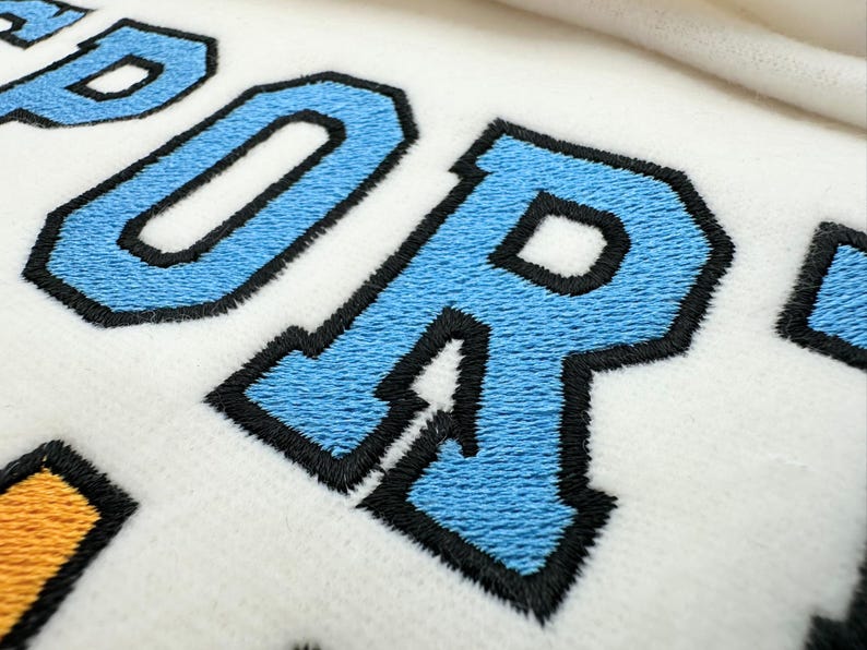 May include: A white fabric surface featuring embroidered lettering with light blue fill and thick black outlines. The visible text includes the letters P, O, and R in a bold, collegiate-style font, with a portion of a yellow embroidered shape visible below.