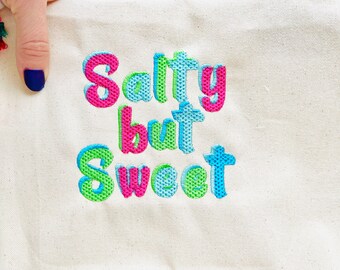 May include: A white canvas tote bag with the embroidered text "Salty but Sweet" in pink, blue, and green.