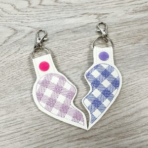 Puede incluir: Dos llaveros en forma de corazón hechos de material blanco con patrones de cuadros vichy bordados. Uno presenta un diseño de cuadros morados con un botón a presión rosa, y el otro tiene un diseño de cuadros azules con un botón a presión morado.