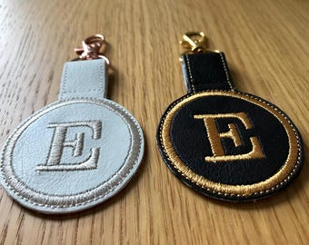 May include: Two leather keychains with a gold embroidered letter E. One keychain is white with a silver ring and the other is black with a gold ring.