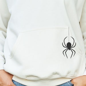 Puede incluir: Una sudadera con capucha blanca con una silueta de araña negra impresa en el bolsillo.