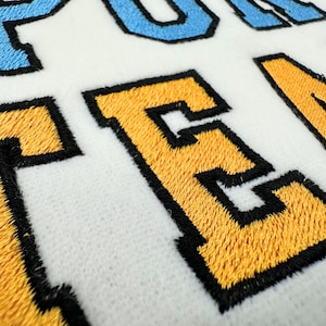 May include: A white fabric surface featuring embroidered lettering with a varsity-style font. The letters are stitched in bright yellow and light blue thread, each outlined with a thick black border, creating a textured and raised appearance.