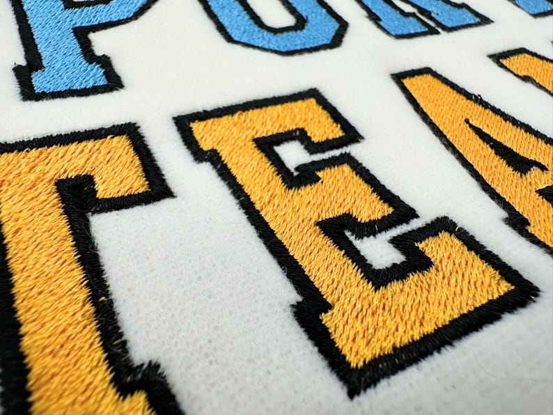 May include: A white fabric surface featuring embroidered lettering with a varsity-style font. The letters are stitched in bright yellow and light blue thread, each outlined with a thick black border, creating a textured and raised appearance.