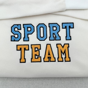 May include: A folded off-white sweatshirt featuring the embroidered text SPORT TEAM in a collegiate font. The word SPORT is stitched in light blue with a black outline, and the word TEAM is stitched in golden yellow with a black outline.