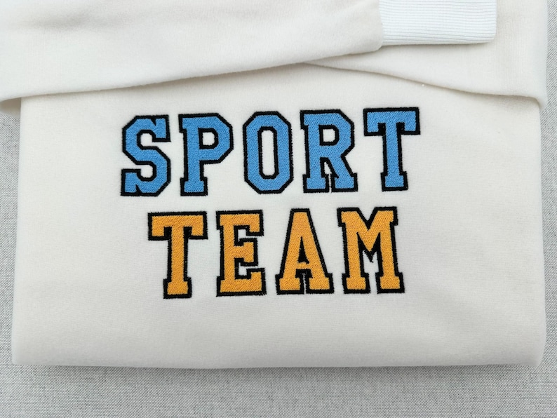 May include: A folded off-white sweatshirt featuring the embroidered text SPORT TEAM in a collegiate font. The word SPORT is stitched in light blue with a black outline, and the word TEAM is stitched in golden yellow with a black outline.